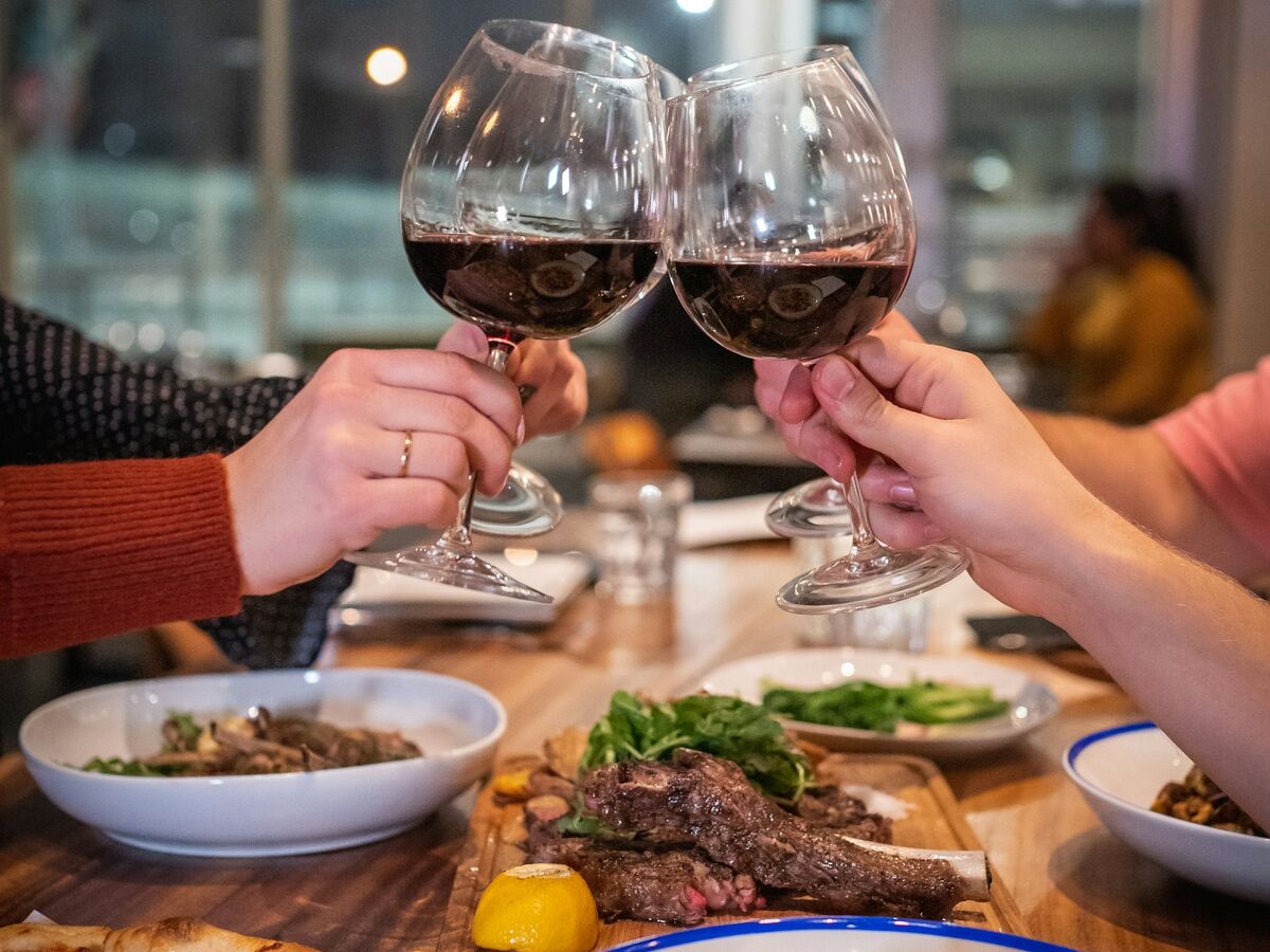 Two people clink glasses of red wine over a table of pasta, pizza and grilled meat.