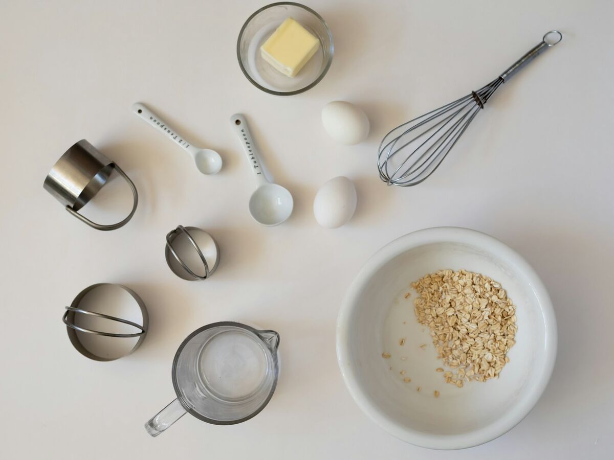 Measuring cups and spoons lie on a white surface with butter, eggs, a whisk and a bowl of oats.