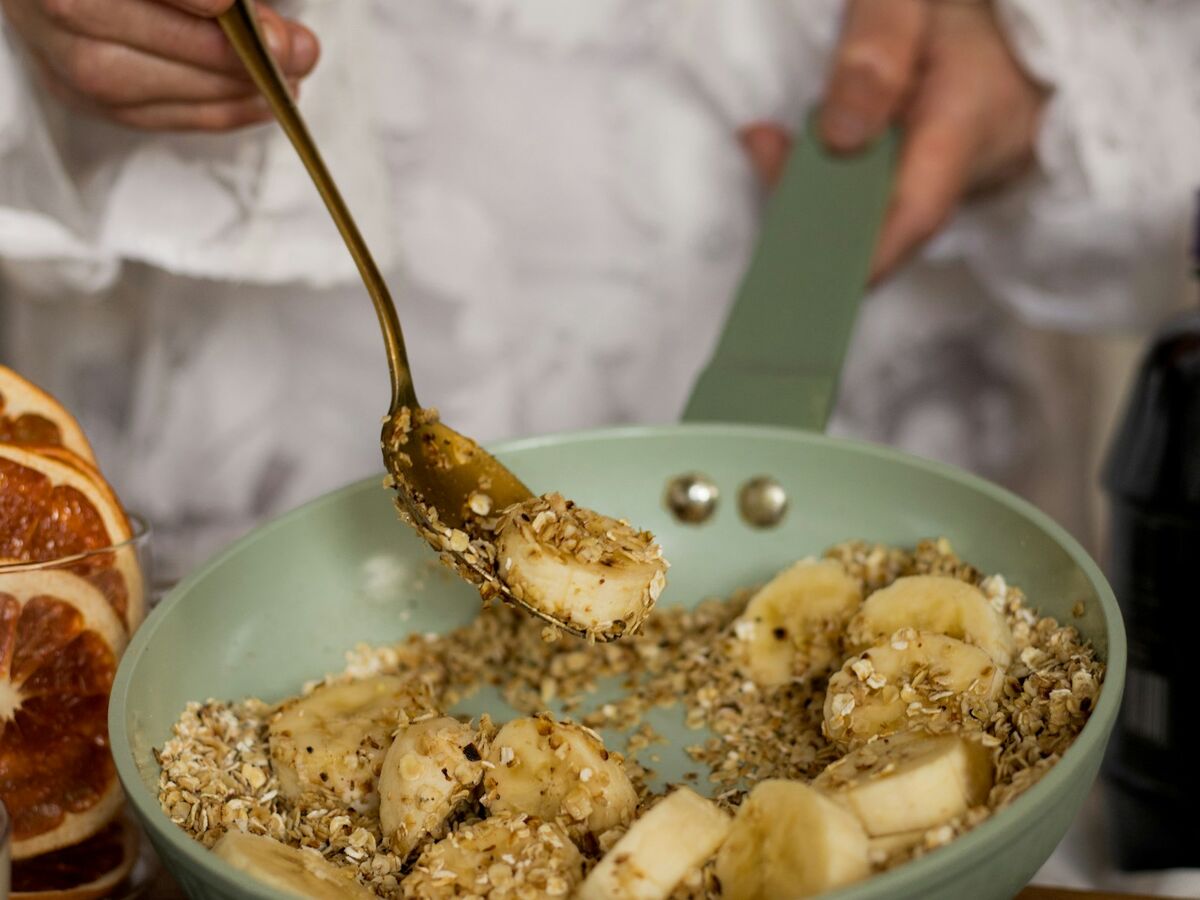 Hands stir sliced banana and oats in a light green pan.
