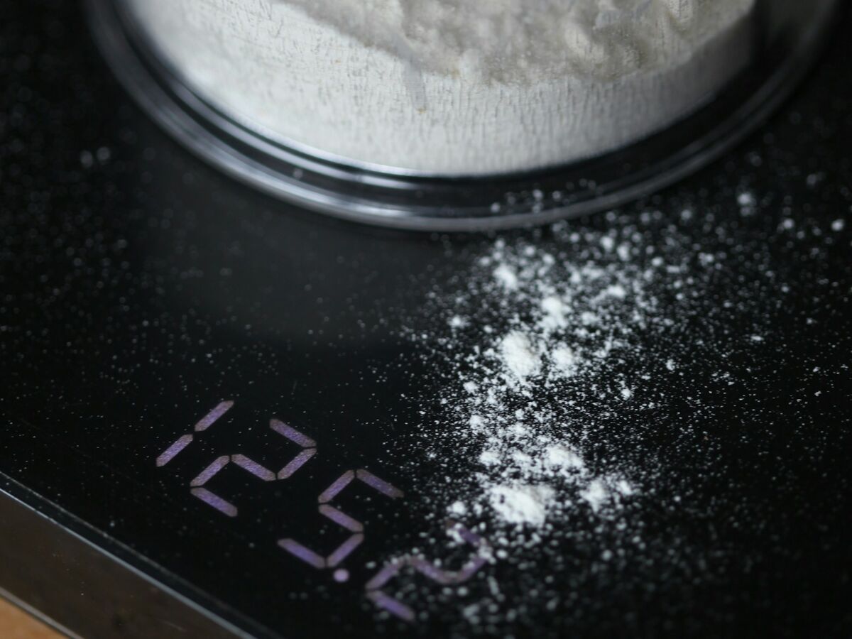 A container of flour sits on a black digital kitchen scale that reads 125.2 with flour dust on the surface.