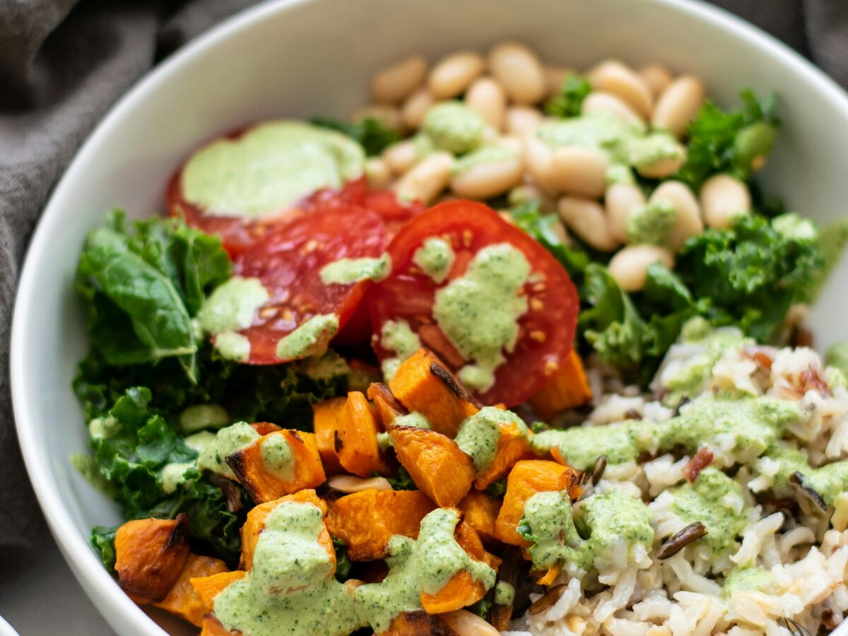 A white bowl holds rice, roasted sweet potato, kale, tomato and white beans topped with green sauce.