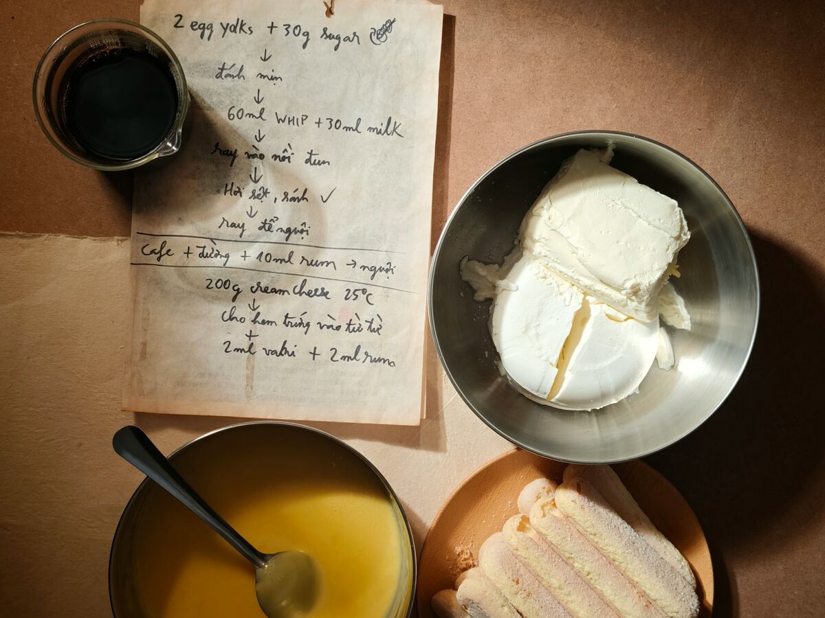 A handwritten recipe card sits on a brown counter beside bowls of mascarpone, custard and ladyfingers.