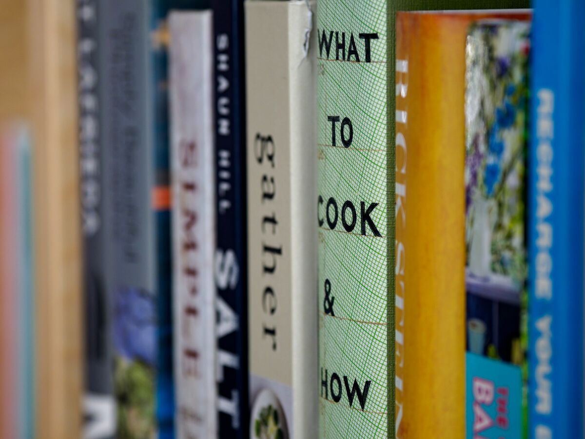 A row of cookbooks stands on a wooden shelf with their spines facing out.