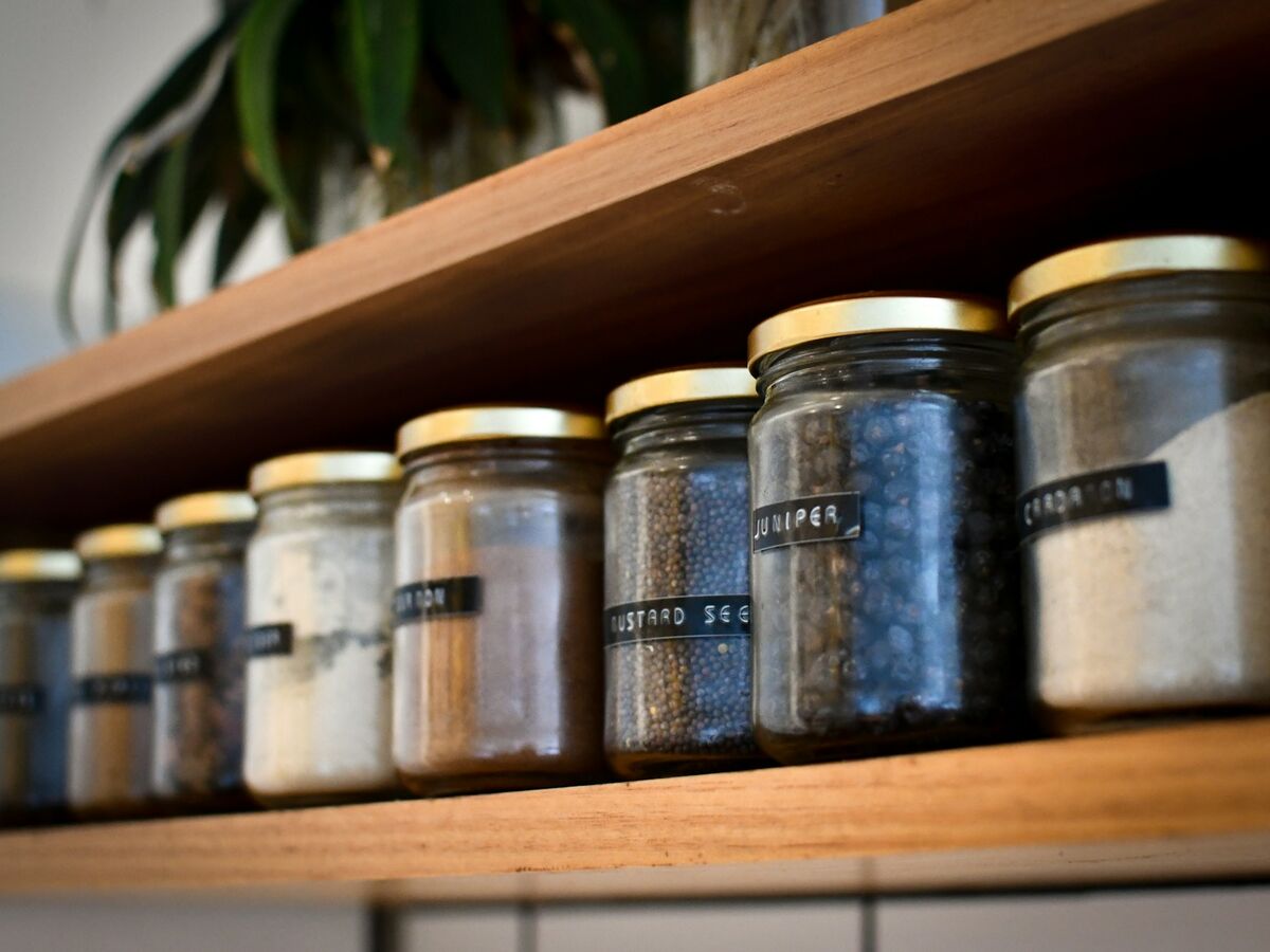 Labeled glass spice jars with gold lids stand in a row on a wooden shelf.