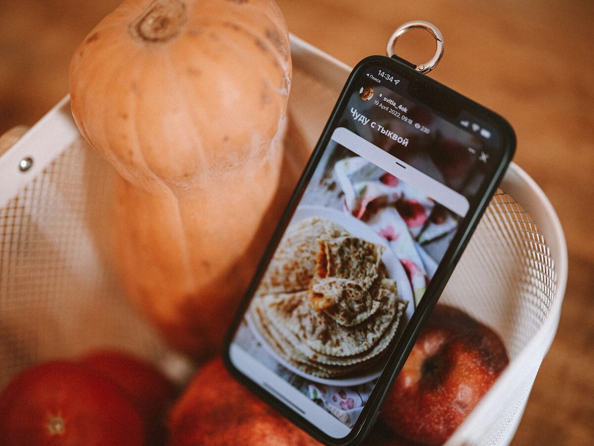 A phone showing a food photo leans in a colander next to a butternut squash and apples.