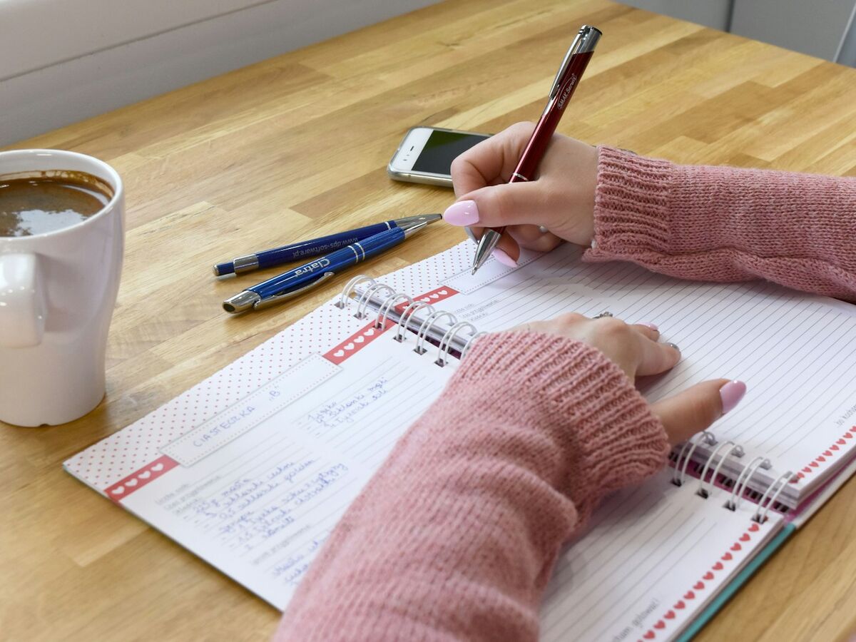 A person in a pink sweater writes in a spiral notebook at a wooden table with coffee and pens.