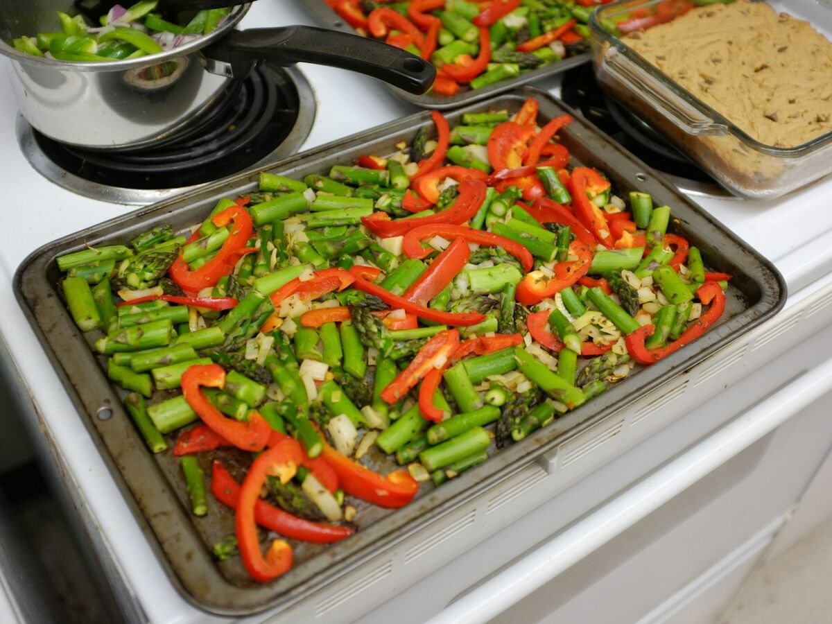 A sheet pan of asparagus and red pepper strips sits on a stovetop next to other pans.